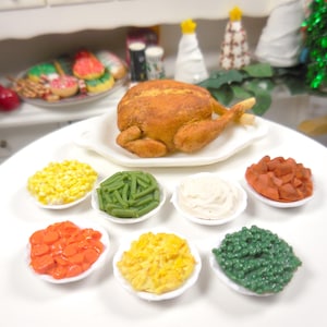 Può includere: Un set da cena del Ringraziamento in miniatura su un tavolo bianco. Il set comprende un tacchino arrosto su un piatto bianco e piccole ciotole di mais, fagiolini, purè di patate, carote e salsa di mirtilli rossi. Decorazioni natalizie sullo sfondo.