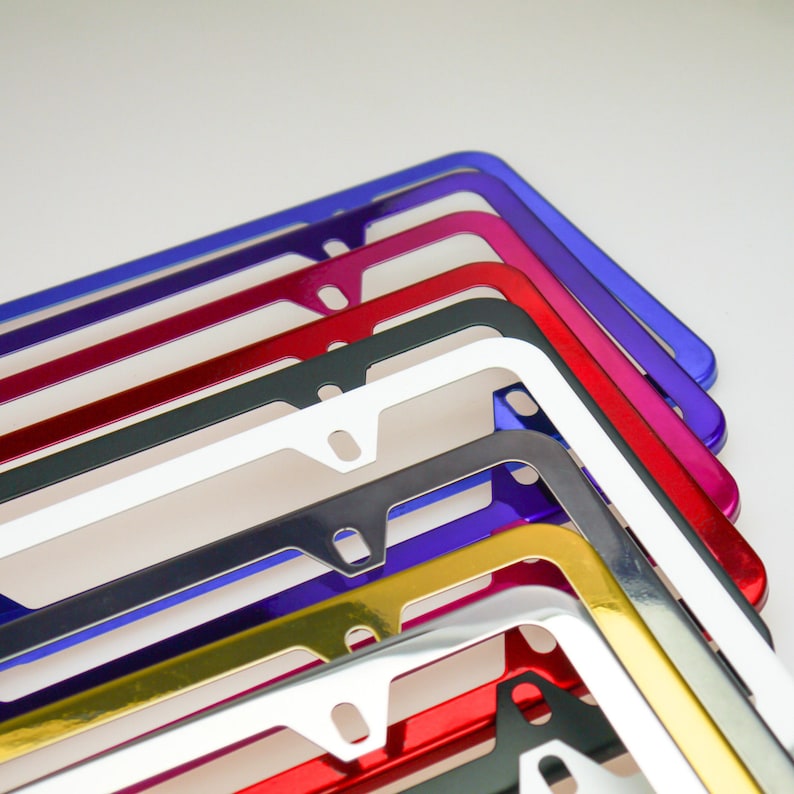May include: A stack of colorful metal license plate frames. The frames are in various colors, including red, gold, silver, black, and blue. The frames are all made of metal and have a shiny finish.
