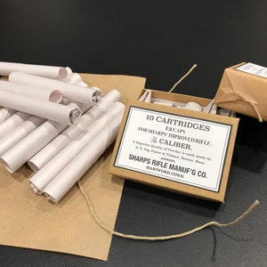 May include: A collection of white paper cartridges and a cardboard box labeled "10 CARTRIDGES FOR SHARPS IMPROVED RIFLE" with additional text. The box is tied with twine, and several cartridges are scattered on a brown paper surface.