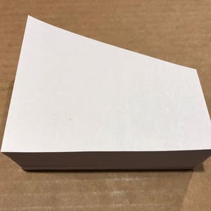 May include: A stack of white paper with a unique angled cut on top. The paper is rectangular with a clean, crisp appearance. The paper is on a brown surface.