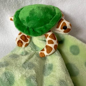 Peut inclure: Une couverture de sécurité en peluche avec une tête et des membres de tortue en peluche attachés à un carré de tissu doux vert clair. La tortue a une carapace vert vif et des membres tachetés de brun. Le tissu est vert clair à pois vert foncé.