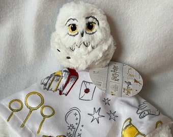 Op de afbeelding: Witte pluche Harry Potter Hedwig veiligheidsdeken met een witte en grijze print van Harry Potter-thema's, waaronder een bril, een sjaal, een toverstaf, een gouden snitch, een brief, een boek, een kwispel en een beker van een Zweinstein-huis. Het label luidt "The Wizarding World Harry Potter".