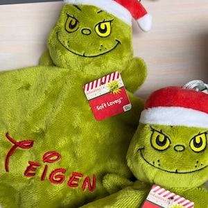 May include: Two green plush Grinch loveys with red Santa hats. Each features an Infantino tag labeled Soft Lovey and a white loop. The left lovey is embroidered with the name Teigen in red, and the right lovey is embroidered with the name Leny in red.