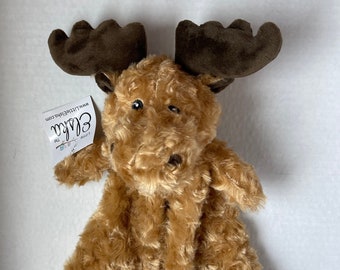 May include: A plush moose toy with brown antlers and a soft, textured body in shades of tan and brown. The toy has small, black eyes and a tag that reads "Elska". The moose is designed as a comfort toy.