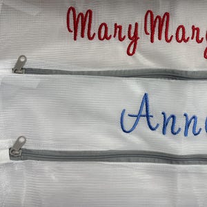 K&ouml;nnte beinhalten: Wei&szlig;e W&auml;schebeutel aus Mesh mit grauen Rei&szlig;verschl&uuml;ssen. Auf dem oberen Beutel ist der Name "Mary Margaret" in roter Schrift gestickt, und auf dem unteren Beutel ist der Name "Anne" in blauer Schrift gestickt. Die Beutel sind f&uuml;r die W&auml;sche bestimmt.