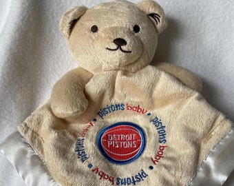 May include: A tan plush baby blanket with a white satin border. The blanket features a brown teddy bear and an embroidered basketball with the words "Detroit Pistons Baby" around it.