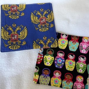 May include: Two square fabric coasters. One coaster is blue with a repeating gold double-headed eagle emblem. The second coaster is black with a repeating pattern of colorful matryoshka dolls featuring floral details and various hair colors.