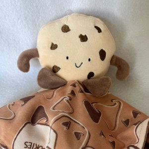 May include: A brown and tan plush cookie blanket with a smiling cookie face attached. The blanket has a pattern of cookies with the word "COOKIES" printed on it.