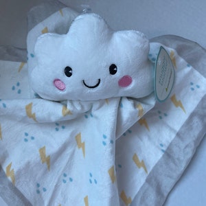 Può includere: Una nuvola di peluche bianca con un viso sorridente e guance rosa è attaccata a una copertina per bambini bianca con un motivo di fulmini gialli e gocce di pioggia blu. La copertina ha un bordo grigio.
