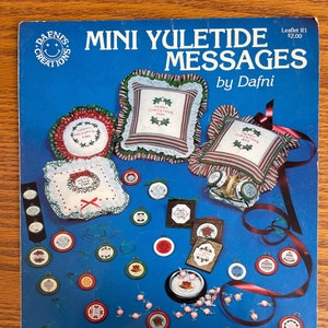 May include: A blue leaflet titled "MINI YULETIDE MESSAGES by Dafni" with various cross-stitch Christmas ornaments and pillows. The ornaments are round with red, green, and white details. The pillows have striped borders and the text "MERRY CHRISTMAS MOM".