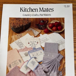 Peut inclure: Ensemble de textiles et d'accessoires de cuisine, comprenant des serviettes, des maniques et un gant de cuisine. Les articles sont dans des tons de rose, de lavande et de blanc, avec des motifs floraux brodés. Le texte "Kitchen Mates" est en haut.