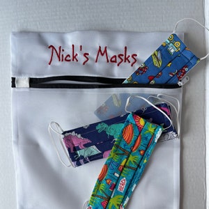 K&ouml;nnte beinhalten: Eine wei&szlig;e Netztasche mit schwarzem Rei&szlig;verschluss und dem rot gestickten Text "Nick's Masks". Die Tasche enth&auml;lt mehrere Gesichtsmasken mit verschiedenen Mustern, darunter Dinosaurier, Autos und Strandszenen.