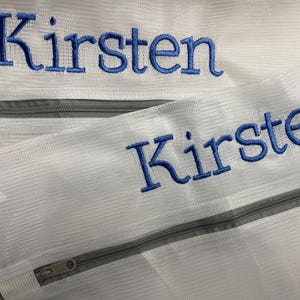 K&ouml;nnte beinhalten: Zwei wei&szlig;e W&auml;schebeutel aus Mesh mit grauen Rei&szlig;verschl&uuml;ssen. Der Name "Kirsten" ist auf jeder Tasche mit blauem Faden gestickt. Die Taschen sind f&uuml;r das Waschen empfindlicher Artikel konzipiert.