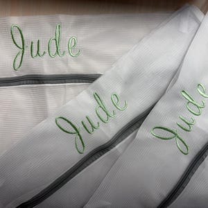 K&ouml;nnte beinhalten: Wei&szlig;e W&auml;schebeutel aus Mesh mit grauen Rei&szlig;verschl&uuml;ssen. Der Name "Jude" ist in gr&uuml;ner Schreibschrift auf jede Tasche gestickt. Die Taschen sch&uuml;tzen Kleidung beim Waschen.
