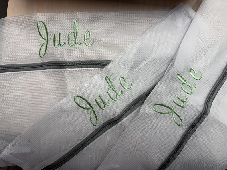 K&ouml;nnte beinhalten: Wei&szlig;e W&auml;schebeutel aus Mesh mit grauen Rei&szlig;verschl&uuml;ssen. Der Name "Jude" ist in gr&uuml;ner Schreibschrift auf jede Tasche gestickt. Die Taschen sch&uuml;tzen Kleidung beim Waschen.
