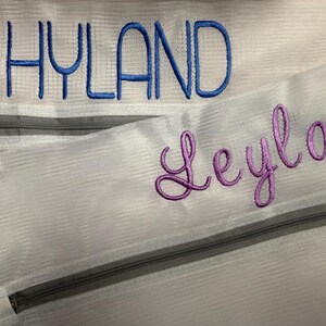 K&ouml;nnte beinhalten: Nahaufnahme von zwei wei&szlig;en Netztaschen mit grauen Rei&szlig;verschl&uuml;ssen. Das Wort "HYLAND" ist in Blau auf die obere Tasche gestickt. Der Name "Leyla" ist in Lila auf die untere Tasche gestickt. Die Taschen sind wahrscheinlich f&uuml;r W&auml;sche oder Aufbewahrung.