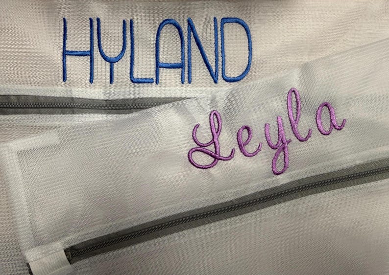 K&ouml;nnte beinhalten: Nahaufnahme von zwei wei&szlig;en Netztaschen mit grauen Rei&szlig;verschl&uuml;ssen. Das Wort "HYLAND" ist in Blau auf die obere Tasche gestickt. Der Name "Leyla" ist in Lila auf die untere Tasche gestickt. Die Taschen sind wahrscheinlich f&uuml;r W&auml;sche oder Aufbewahrung.