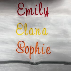 K&ouml;nnte beinhalten: Wei&szlig;er W&auml;schebeutel aus Mesh mit drei gestickten Namen: "Emily" in Rot, "Elana" in Gelb und "Sophie" in Orange. Die Tasche hat einen grauen Rei&szlig;verschluss am unteren Rand.
