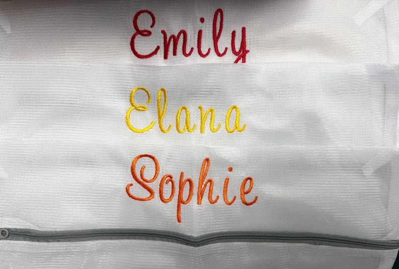 K&ouml;nnte beinhalten: Wei&szlig;er W&auml;schebeutel aus Mesh mit drei gestickten Namen: "Emily" in Rot, "Elana" in Gelb und "Sophie" in Orange. Die Tasche hat einen grauen Rei&szlig;verschluss am unteren Rand.