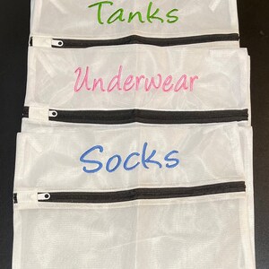 K&ouml;nnte beinhalten: Wei&szlig;e W&auml;schebeutel aus Mesh mit schwarzen Rei&szlig;verschl&uuml;ssen. Jeder Beutel hat gestickten Text: "Tanks" in Gr&uuml;n, "Underwear" in Pink und "Socks" in Blau. Die Beutel sind zum Organisieren der W&auml;sche gedacht.