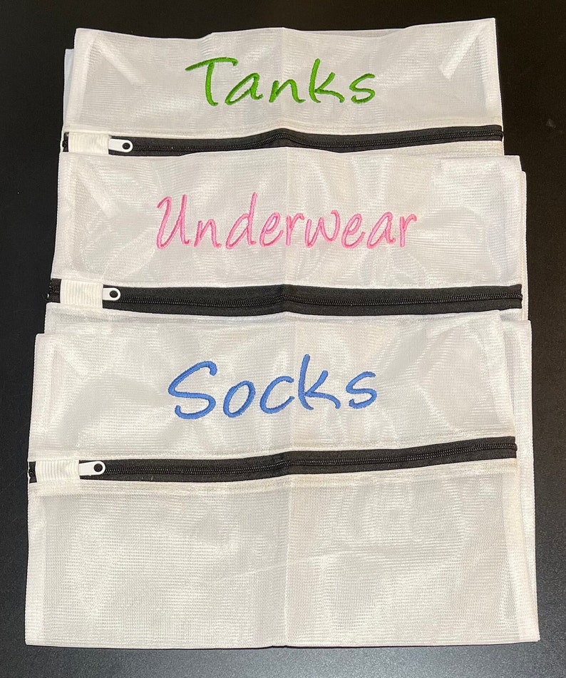 K&ouml;nnte beinhalten: Wei&szlig;e W&auml;schebeutel aus Mesh mit schwarzen Rei&szlig;verschl&uuml;ssen. Jeder Beutel hat gestickten Text: "Tanks" in Gr&uuml;n, "Underwear" in Pink und "Socks" in Blau. Die Beutel sind zum Organisieren der W&auml;sche gedacht.