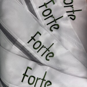 K&ouml;nnte beinhalten: Nahaufnahme mehrerer wei&szlig;er Stoffstreifen mit dem Wort "Forte" in Gr&uuml;n gestickt. Der Stoff hat ein strukturiertes, geripptes Aussehen, und jeder Streifen hat einen grauen Rei&szlig;verschluss. Das Wort "Forte" wird mehrfach wiederholt.