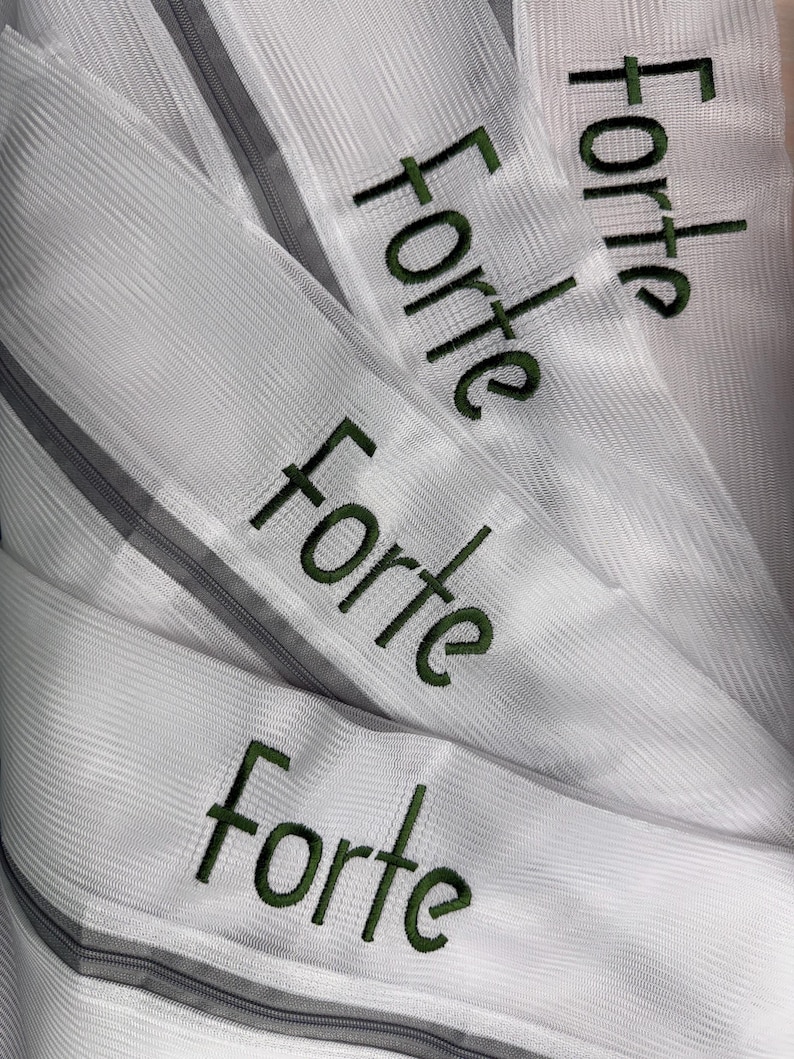 K&ouml;nnte beinhalten: Nahaufnahme mehrerer wei&szlig;er Stoffstreifen mit dem Wort "Forte" in Gr&uuml;n gestickt. Der Stoff hat ein strukturiertes, geripptes Aussehen, und jeder Streifen hat einen grauen Rei&szlig;verschluss. Das Wort "Forte" wird mehrfach wiederholt.