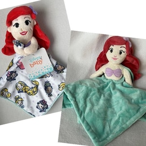 Może przedstawiać: Dwie pluszowe lalki Ariel z Disneya, syrenki. Jedna lalka trzyma fioletową muszlę i leży na białym kocu z logo Disney Baby i obrazkami innych księżniczek Disneya. Druga lalka leży na zielonym kocu.