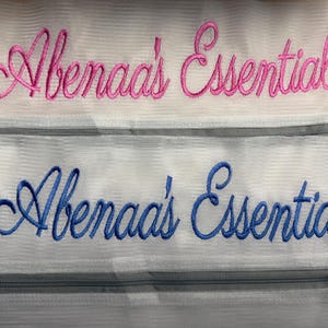 K&ouml;nnte beinhalten: Zwei wei&szlig;e Netztaschen mit gesticktem Text. Die obere Tasche hat einen rosa Schriftzug in Schreibschrift mit der Aufschrift "Abenaa's Essentials". Die untere Tasche hat einen blauen Schriftzug in Schreibschrift mit der Aufschrift "Abenaa's Essentials".