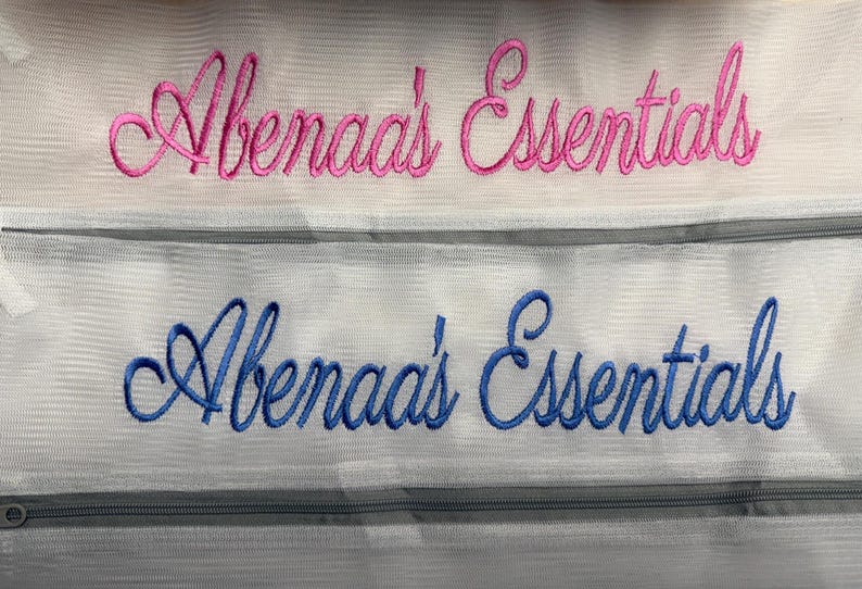 K&ouml;nnte beinhalten: Zwei wei&szlig;e Netztaschen mit gesticktem Text. Die obere Tasche hat einen rosa Schriftzug in Schreibschrift mit der Aufschrift "Abenaa's Essentials". Die untere Tasche hat einen blauen Schriftzug in Schreibschrift mit der Aufschrift "Abenaa's Essentials".