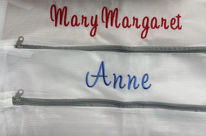 K&ouml;nnte beinhalten: Wei&szlig;e W&auml;schebeutel aus Mesh mit grauen Rei&szlig;verschl&uuml;ssen. Auf dem oberen Beutel ist der Name "Mary Margaret" in roter Schrift gestickt, und auf dem unteren Beutel ist der Name "Anne" in blauer Schrift gestickt. Die Beutel sind f&uuml;r die W&auml;sche bestimmt.