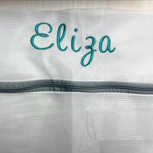 K&ouml;nnte beinhalten: Wei&szlig;er W&auml;schebeutel aus Mesh mit dem t&uuml;rkis gestickten Namen "Eliza". Die Tasche hat einen grauen Rei&szlig;verschluss. Das Mesh-Material ist sichtbar, und das Design ist schlicht und funktional.