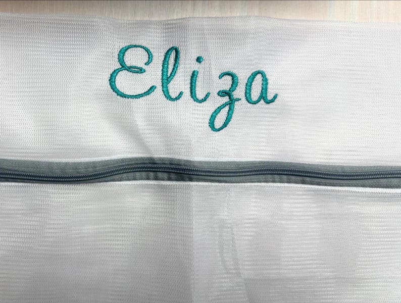 K&ouml;nnte beinhalten: Wei&szlig;er W&auml;schebeutel aus Mesh mit dem t&uuml;rkis gestickten Namen "Eliza". Die Tasche hat einen grauen Rei&szlig;verschluss. Das Mesh-Material ist sichtbar, und das Design ist schlicht und funktional.