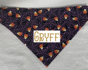 May include: Triangular dog bandana with a Halloween theme. The fabric is black with a purple spiderweb pattern and candy corn accents. A white rectangle with the name "GRYFF" in gold lettering is centered on the bandana.
