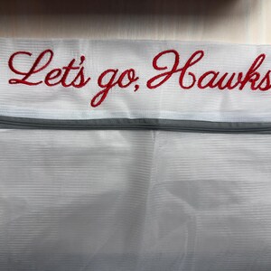 K&ouml;nnte beinhalten: Wei&szlig;er W&auml;schebeutel aus Mesh mit grauem Rei&szlig;verschluss und dem gestickten Text "Let's go, Hawks!" in roter Schrift. Die Tasche ist f&uuml;r das Waschen empfindlicher Artikel konzipiert und hat einen Rei&szlig;verschluss.