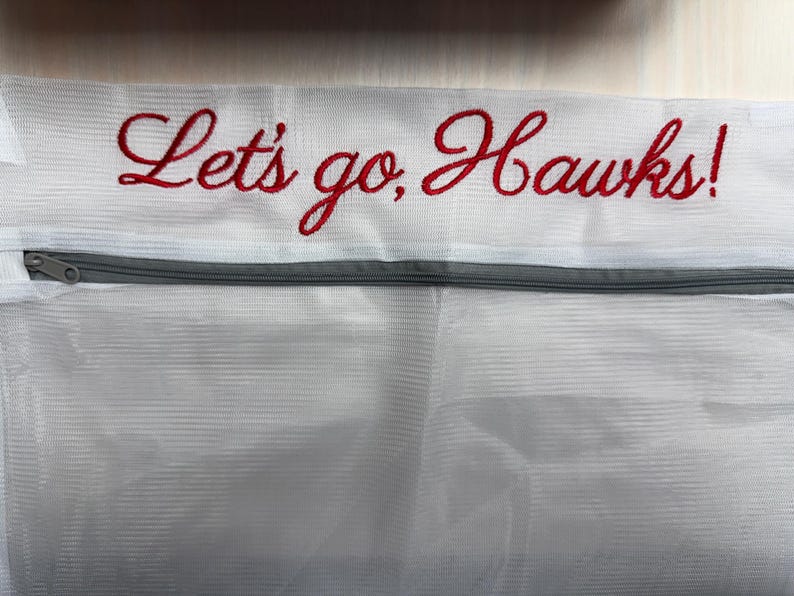 K&ouml;nnte beinhalten: Wei&szlig;er W&auml;schebeutel aus Mesh mit grauem Rei&szlig;verschluss und dem gestickten Text "Let's go, Hawks!" in roter Schrift. Die Tasche ist f&uuml;r das Waschen empfindlicher Artikel konzipiert und hat einen Rei&szlig;verschluss.
