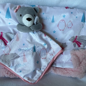 May include: A grey and white baby blanket with a grey teddy bear attached. The blanket has a pink border and features a winter theme with penguins, trees, and stars.
