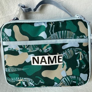 May include: A green and beige lunch bag with a dinosaur skeleton pattern. The bag has a light blue zipper and a light blue strap. The bag has a white label with the word "NAME" on it.
