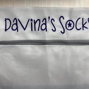 K&ouml;nnte beinhalten: Wei&szlig;er W&auml;schebeutel aus Mesh mit einem grauen Rei&szlig;verschluss. Der Beutel hat den gestickten Text "Davina's Socks" in Dunkelblau, mit einem Sternsymbol. Zum Waschen von Socken.