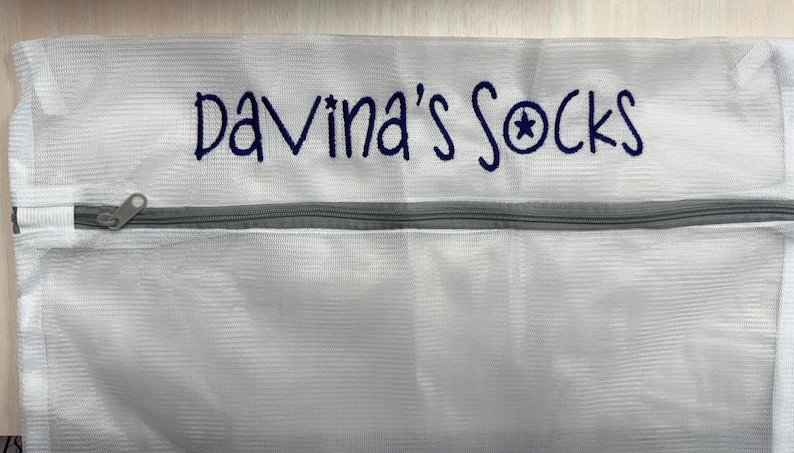 K&ouml;nnte beinhalten: Wei&szlig;er W&auml;schebeutel aus Mesh mit einem grauen Rei&szlig;verschluss. Der Beutel hat den gestickten Text "Davina's Socks" in Dunkelblau, mit einem Sternsymbol. Zum Waschen von Socken.