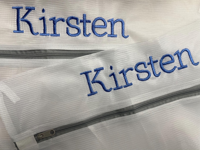 K&ouml;nnte beinhalten: Zwei wei&szlig;e W&auml;schebeutel aus Mesh mit grauen Rei&szlig;verschl&uuml;ssen. Der Name "Kirsten" ist auf jeder Tasche mit blauem Faden gestickt. Die Taschen sind f&uuml;r das Waschen empfindlicher Artikel konzipiert.