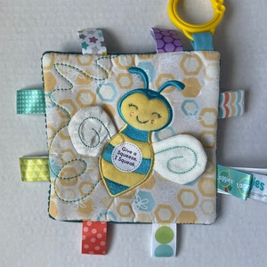 May include: A yellow and blue bee with a honeycomb pattern on a soft, square, multi-textured baby toy. The bee has a smiling face and says "Give a Squeeze, I Squeak." The toy has a variety of colorful ribbons and a yellow plastic ring for attaching to a stroller or car seat.