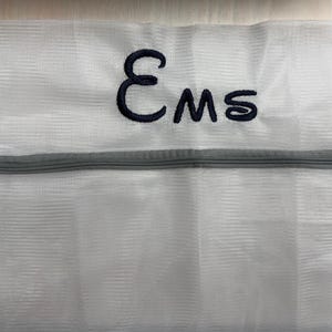 K&ouml;nnte beinhalten: Wei&szlig;er W&auml;schebeutel aus Mesh mit grauem Rei&szlig;verschluss. Der Beutel hat die gestickten Buchstaben "Ems" in Marineblau. Der Beutel dient zum Schutz empfindlicher Kleidung beim Waschen.