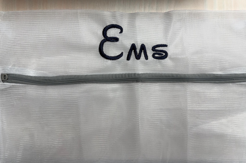 K&ouml;nnte beinhalten: Wei&szlig;er W&auml;schebeutel aus Mesh mit grauem Rei&szlig;verschluss. Der Beutel hat die gestickten Buchstaben "Ems" in Marineblau. Der Beutel dient zum Schutz empfindlicher Kleidung beim Waschen.