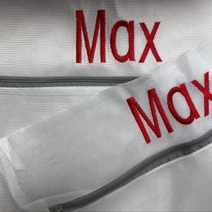 K&ouml;nnte beinhalten: Nahaufnahme von zwei wei&szlig;en W&auml;schebeuteln aus Mesh mit grauen Rei&szlig;verschl&uuml;ssen. Der Name "Max" ist rot auf beiden Beuteln gestickt. Die Beutel sind f&uuml;r die W&auml;sche empfindlicher Artikel gedacht.