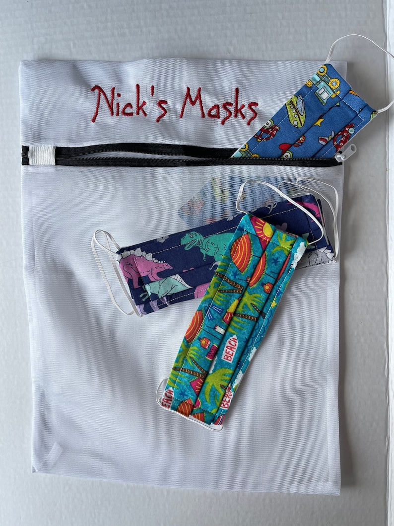 K&ouml;nnte beinhalten: Eine wei&szlig;e Netztasche mit schwarzem Rei&szlig;verschluss und dem rot gestickten Text "Nick's Masks". Die Tasche enth&auml;lt mehrere Gesichtsmasken mit verschiedenen Mustern, darunter Dinosaurier, Autos und Strandszenen.