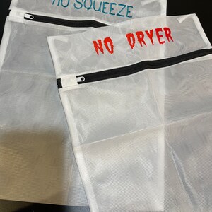 K&ouml;nnte beinhalten: Drei wei&szlig;e W&auml;schebeutel aus Netzstoff mit schwarzen Rei&szlig;verschl&uuml;ssen. Auf dem oberen Beutel steht "NO SQUEEZE" in T&uuml;rkis gestickt, auf dem unteren "NO DRYER" in Rot. Die Beutel sch&uuml;tzen empfindliche W&auml;sche.