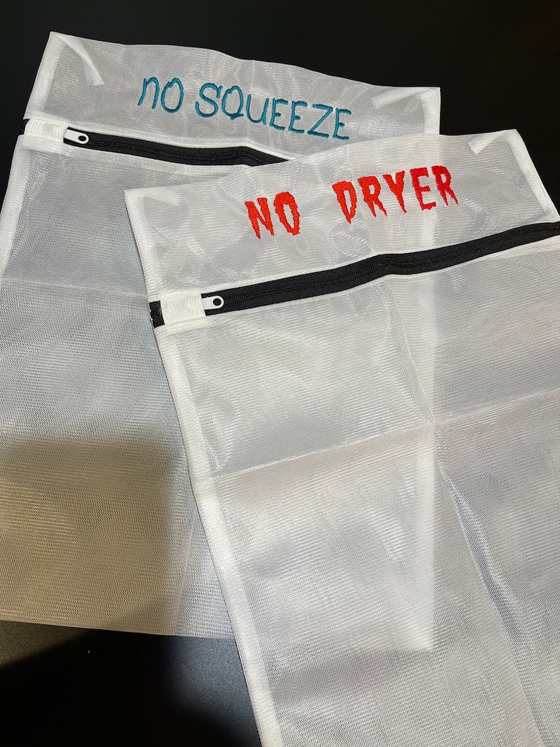 K&ouml;nnte beinhalten: Drei wei&szlig;e W&auml;schebeutel aus Netzstoff mit schwarzen Rei&szlig;verschl&uuml;ssen. Auf dem oberen Beutel steht "NO SQUEEZE" in T&uuml;rkis gestickt, auf dem unteren "NO DRYER" in Rot. Die Beutel sch&uuml;tzen empfindliche W&auml;sche.