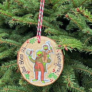 May include: A wooden Christmas ornament featuring a green frog wearing a top hat and a red scarf, holding a book that says "God bless us, everyone".