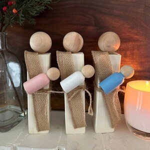 May include: Three wooden figures with painted heads and bodies, standing on white blocks wrapped in burlap ribbon. The figures are holding smaller wooden figures painted pink, white, and blue.